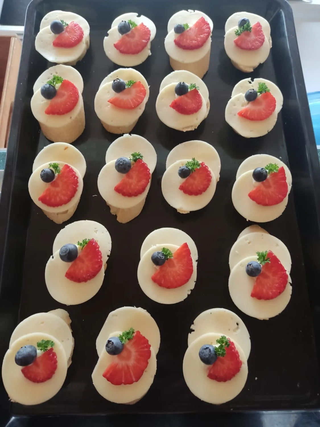 Thörnich – EventCatering – Ein Tablett mit Crostini-Häppchen. Die sichtbaren Zutaten lassen darauf schließen, dass es sich um eine Art Crostini handelt, die mit Ziegenkäse, Brombeeren und anderen Garnituren wie Blaubeeren oder kleinen grünen Kräutern garniert sind.