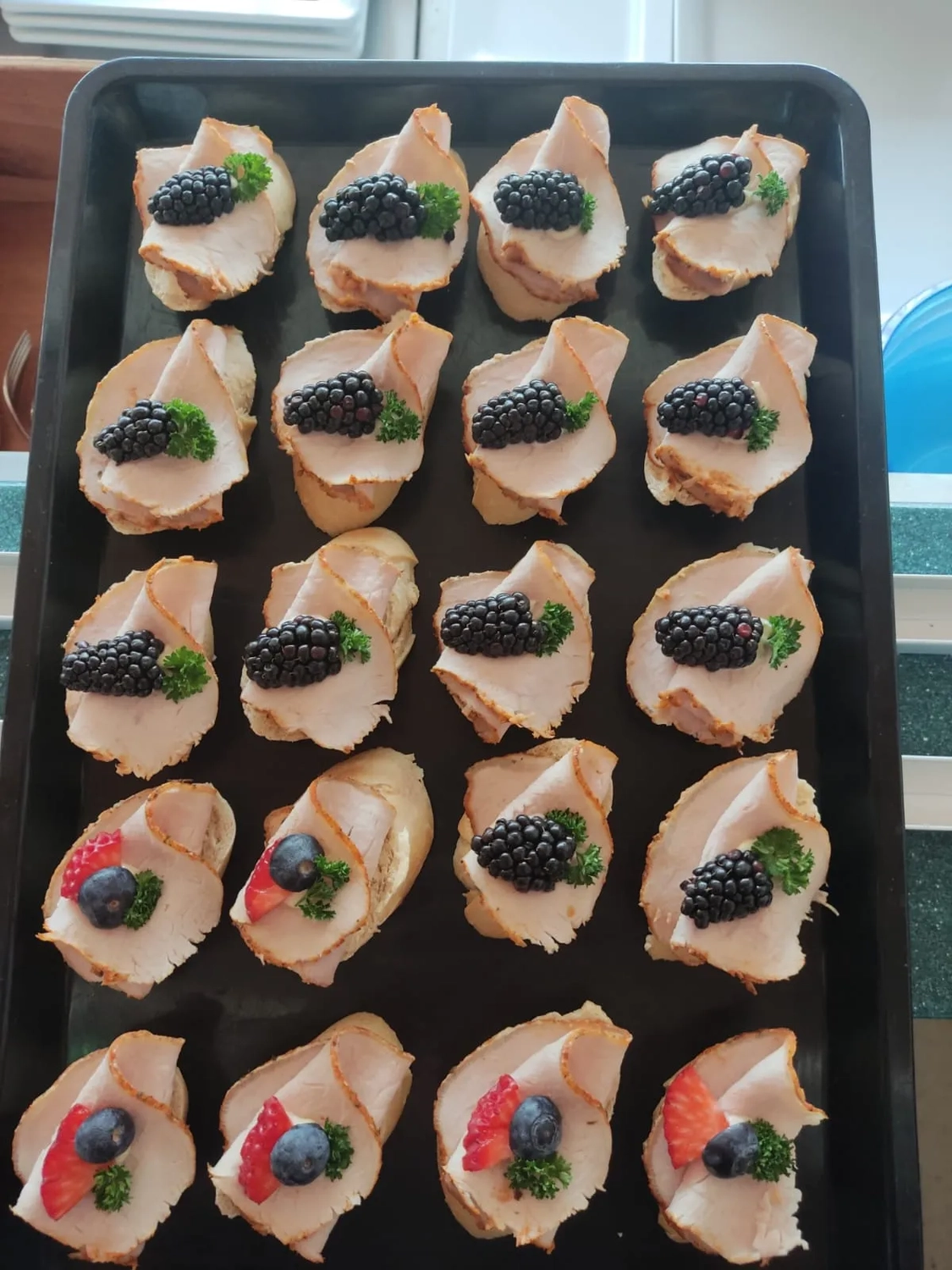 Thörnich – EventCatering – Ein Tablett mit Crostini-Häppchen. Die sichtbaren Zutaten lassen darauf schließen, dass es sich um eine Art Crostini handelt, die mit Ziegenkäse, Brombeeren und anderen Garnituren wie Blaubeeren oder kleinen grünen Kräutern garniert sind.